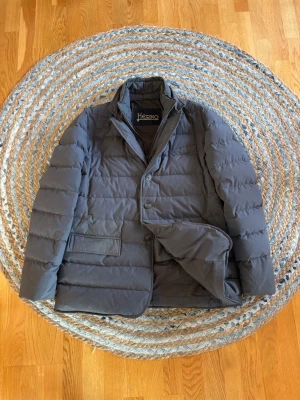 Mörkgrå pufferjacka från Herno - Stilren mörkgrå pufferjacka från Herno med klassisk krage och knäppning framtill. Jackan har två stora fickor med lock och quiltad design som ger en modern look. Perfekt för kalla vinterdagar och håller dig varm med sin vadderade insida. Observera att det finns 2 fläckar på fickorna, inget som syns vid användning 