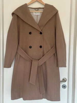 Beige ullrock med huva från Happy Holly - Stilren beige ullrock från Happy Holly med stor huva, knytskärp i midjan och dubbelknäppning framtill. Rocken har långa ärmar och en klassisk, rak passform som ger en snygg siluett. Perfekt för kyliga dagar när du vill vara både varm och trendig.