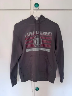 Grå Saint L hoodie - Säljer en grå hoodie från Saint L med tryck i vitt och vinrött framtill. Hoodien har huva med dragsko och en stor magficka. Materialet är mjukt och skönt, perfekt för en avslappnad stil. Snygg logga och coola detaljer gör den till ett måste i garderoben. Perfekt för dig som vill ha en niche tröja som är as snygg!