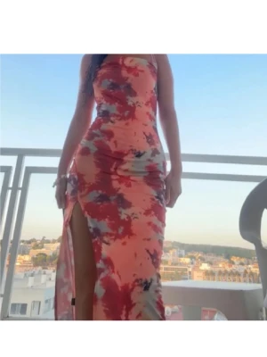 Sommar klänning - Säljer en lång klänning med tunna axelband och snygg slits. Klänningen har ett färgstarkt batikmönster i rött, rosa, grått och beige. Den är figurnära och har en draperad urringning som ger en trendig vibe. Perfekt för sommarkvällar och strandpromenader. Passar s också. Köpt i mallorca. Endast använd 1 gång.