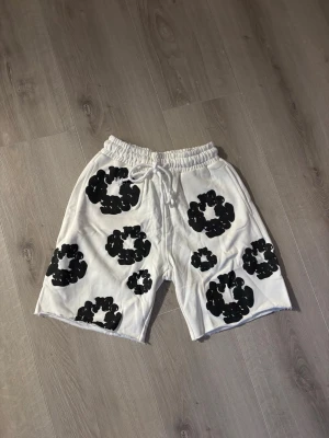 Vita shorts från Denim Tears M - Säljer ett par vita shorts från Denim Tears med svart blommigt tryck. Shortsen har resårmidja med snörning och är tillverkade i mjuk bomull. Perfekta för sommaren och har en avslappnad passform. Pris kan diskuteras vid snabb affär 😊