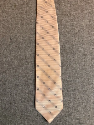 Beige slips med rutmönster La Ploma Italy - Stilren beige slips från La Ploma med diskret rutmönster i mörkblått. Slipsen har en lätt glansig yta och tunna horisontella ränder som ger extra struktur. Perfekt accessoar för att lyfta din outfit med en elegant touch.