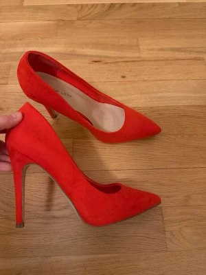 Orangea pumps från New Look - Snygga orangea pumps från New Look med spetsig tå och hög klack. Skorna är klädda i mjuk mockaimitation som ger en lyxig känsla och har en klassisk siluett som passar till många olika stilar. Perfekta för dig som vill sticka ut med färg.