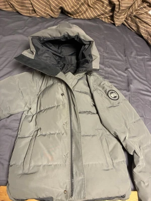 Grå dunjacka från Canada Goose - Snygg grå dunjacka från Canada Goose med huva och klassisk logotyp på ärmen. Jackan har dragkedja och knappar framtill samt två stora fickor. Perfekt för kalla vinterdagar tack vare det varma dunfodret.