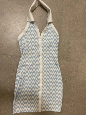 Blåvit halterneckklänning med vågmönster - Säljer en kort halterneckklänning i blått och vitt med vågigt mönster och knappar framtill. Klänningen har en krage och är ärmlös, perfekt för sommarens alla tillfällen. Materialet känns mjukt och svalt. Kan mötts upp i Stockholm.