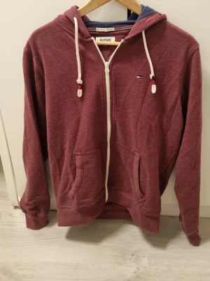 Vinröd hoodie från Tommy Hilfiger - Snygg vinröd hoodie från Tommy Hilfiger med dragkedja, vita snören och liten logga på bröstet. Mjuk bomullsblandning, kängurufickor och fodrad huva med blå insida. Perfekt för en avslappnad och stilren look.