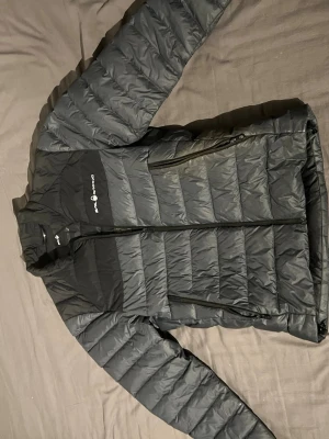 Svart dunjacka från Sail Racing - Snygg svart dunjacka från Sail Racing med diskret logga på bröstet. Jackan har två stora dragkedjeförsedda fickor framtill och quiltad design. Perfekt för kalla vinterdagar och har en modern, sportig look. Som ny