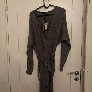 NY !Grå klänning med bälte - Mysig grå klänning i ribbat material med långa ärmar och knytskärp i midjan. Djup V-ringning och avslappnad passform. Enkel och stilren design som passar de flesta stilar. Ny med prislapp