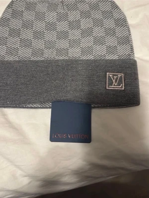 Grå Louis Vuitton mössa - Snygg grå mössa från Louis Vuitton med klassiskt rutmönster och vikt kant. LV-logga broderad framtill. Tillverkad i mjukt material som känns lyxigt och bekvämt. Perfekt accessoar för att höja din stil under kalla dagar.