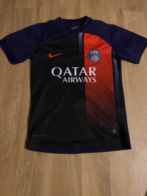 PSG matchtröja Hakimi #2 Nike - Säljer en officiell Paris Saint-Germain matchtröja från Nike med Hakimi och nummer 2 på ryggen. Tröjan är svart och mörkblå med röd detalj och Qatar Airways-logga på bröstet. Perfekt för fotbollsfans som vill visa sitt stöd för PSG.