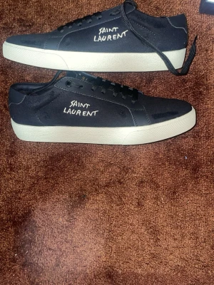 Saint Laurent svarta låga sneakers - Svarta sneakers från Saint Laurent med vit sula och handskriven logga på sidan. Skorna har klassisk låg profil, svarta skosnören och är tillverkade i canvasmaterial. Perfekta för en clean och stilren look. Kvitto finns som äkta bevis 