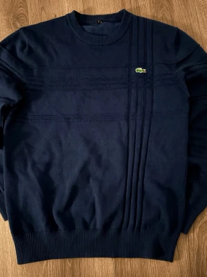 Mörkblå sweatshirt från Lacoste - Snygg mörkblå tröja från Lacoste med diskreta ribbade detaljer och det klassiska krokodilmärket på bröstet. Tröjan har rund halsringning och ribbade muddar vid ärmslut och nederkant. Perfekt för en stilren och avslappnad look.