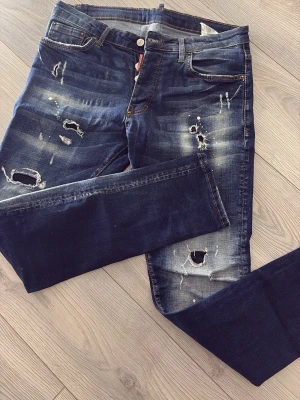 Mörkblå slitna jeans från Dsquared2 - Säljer ett par mörkblå jeans från Dsquared2 med tydliga slitningar och hål för en riktigt edgy look. Jeansen har skinny fit, klassiska fem fickor och kontrastsömmar. Tillverkade i Italien och perfekta för dig som gillar streetstyle.