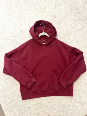 Vinröd hoodie från Gina tricot  - Säljer en enkel och stilren vinröd hoodie med huva och stor magficka framtill. Hoodien har ribbade muddar vid ärmslut och nederkant. Perfekt för dig som gillar en clean look och vill ha något bekvämt att slänga på sig. Materialet känns mjukt och skönt.