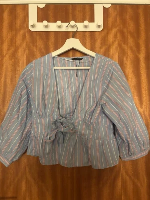 Blå randig blus med knytband från Zara - Söt blus i ljusblått med vita och rosa ränder. Djup v-ringning och knytband i midjan ger en feminin siluett. Blusen har trekvartsärm och lätt puffiga ärmar. Tillverkad i ett tunt, vävt material som känns luftigt och skönt.I storlek M men passar mig som har vanligvis S/Xs