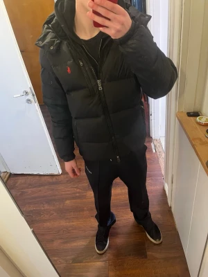 Svart dunjacka från Polo Ralph Lauren - Säljer en svart dunjacka från Polo Ralph Lauren med röd logga på bröstet. Jackan har huva med dragsko, flera dragkedjeförsedda fickor och en puffig, varm design. Perfekt för kalla dagar och riktigt snygg streetstil. Självklart äkta Skicket ser ni på bilderna hör av er vid frågor