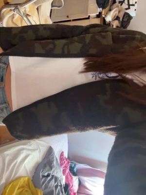 Camo-mönstrad kofta i grönt - Säljer en camo-mönstrad kofta i olika nyanser av grönt och svart. Koftan har en rak passform, långa ärmar och är öppen framtill utan knappar eller dragkedja. Perfekt för dig som gillar streetwear och vill ha en chill look.