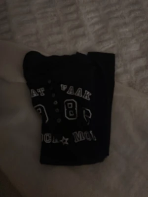 Svart långärmad topp med tryck - Cool svart långärmad topp med vitt tryck framtill där det står 'WAAT FAAK 1986 ROCK MOI'. Toppen har knappar vid halsen och är figurnära i modellen. Perfekt för dig som gillar streetstyle och vill ha en unik detalj i garderoben.