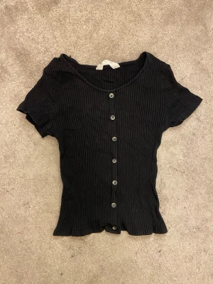 Svart ribbad topp från H&M - Säljer en svart ribbad topp från H&M med korta ärmar och knappar framtill. Toppen har en figurnära passform och är tillverkad i mjukt material som känns skönt mot huden. 