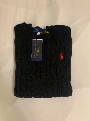 Svart kabelstickad tröja Polo Ralph Lauren - Kabelstickad svart tröja från Polo Ralph Lauren med rund halsringning och röd broderad logga på bröstet. Tröjan har långa ärmar och ribbade muddar vid ärmslut och nederkant. Klassisk och stilren design som passar till mycket.mått: längd: 60cm. Axelbredd: 35cm