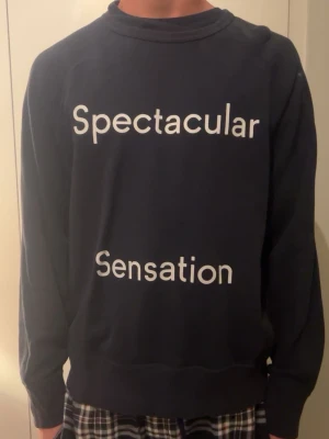 Mörkblå sweatshirt med tryck / Modellen är 182 - Mörkblå sweatshirt med vit text på framsidan där det står 'Spectacular Sensation'. Tröjan har rund halsringning och långa ärmar. Perfekt för en avslappnad och trendig stil. Nypris 6000kr/Mitt pris- 1600kr. Säljer eftersom att jag rensar garderoben.