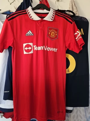Manchester United matchtröja Adidas - Röd Manchester United matchtröja från Adidas med vita detaljer och klubbmärke på bröstet. Tröjan har vit krage med röda inslag, svarta axelränder och TeamViewer-logga framtill. Tillverkad i lätt funktionsmaterial, perfekt för fotboll eller träning.