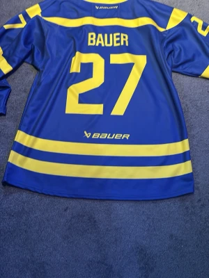 Bauer authentic collection limted edition  - Säljer en blå hockeytröja från Bauer extremt limted upplaga Authentic Collection med gula detaljer och nummer 27 på ryggen. Tröjan har långa ärmar, gula ränder och är tillverkad i 100% polyester. Perfekt för hockeyträning eller som supporterplagg.