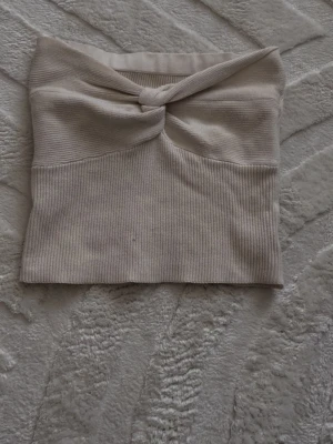 Topp - Trendig ljus beige bandeau tubtopp med twistad detalj framtill. Toppen är ribbad och axelbandslös, perfekt för att styla med höga jeans eller kjol. Mjuk och stretchig för en bekväm passform.