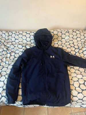 Marinblå jacka från Under Armour - Snygg marinblå hoodie från Under Armour med dragkedja framtill och justerbar huva. Hoodien har två fickor på sidorna och ett vitt Under Armour-logotyptryck på bröstet. Tillverkad i ett mjukt och bekvämt material som passar perfekt till vardags.