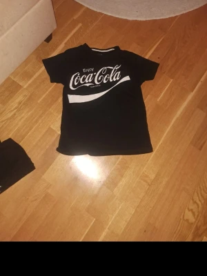 Svart Coca-Cola t-shirt - Säljer en svart t-shirt från Coca-Cola med klassiskt vitt tryck framtill. T-shirten har rund hals och korta ärmar, perfekt för dig som gillar retro och streetwear vibes. Materialet är mjuk bomull som känns skönt mot huden.