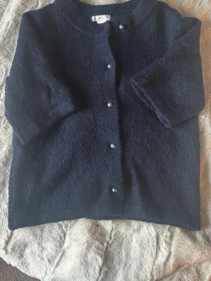 Svart kofta med pärlknappar - Mysig svart kofta med rund halsringning och pärlknappar framtill. Koftan har en rak passform och trekvartsärmar. Materialet ser ut att vara mjukt och fluffigt, perfekt för lager på lager. Enkel och stilren design som passar till det mesta.