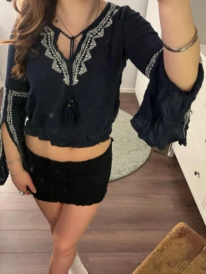 Svart blus med broderi och vida ärmar - Säljer en svart blus med vita broderade detaljer och knytband i halsen ifrån Hollister . Blusen har vida, fladdriga ärmar och croppad passform som ger en bohemisk vibe. Perfekt till sommaren eller festivalen!