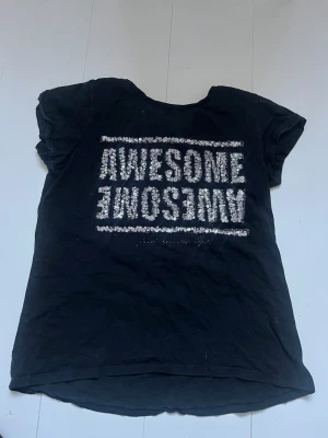 Svart t-shirt med text från KappAhl - Cool svart t-shirt från KappAhl med vit glittrig texten 'AWESOME' tryckt två gånger på framsidan. Klassisk rund halsringning och korta ärmar. Tillverkad i mjuk bomull som är skön att bära.