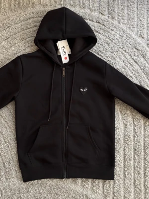 Svart hoodie från Comme des Garçons Play - Svart hoodie med dragkedja från Comme des Garçons Play. Har huva med dragsko, två fickor fram och det ikoniska hjärta-med-ögon-märket på bröstet samt en stor version på ryggen. Tillverkad i mjukt bomullsmaterial för en clean och stilren look. Helt ny med lappen kvar 