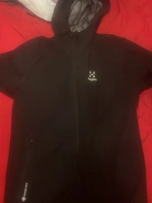 Svart Haglöfs regnjacka med huva - Snygg svart regnjacka från Haglöfs med huva och dragkedja framtill. Tillverkad i slitstarkt Gore-Tex-material som står emot regn och vind. Jackan har diskret logga på bröstet och praktiska fickor. Perfekt för blöta dagar när du vill hålla dig torr.