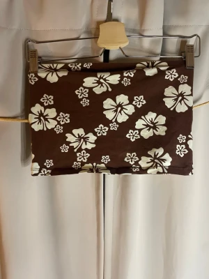 Brun blommig bandeau tubtopp - Säljer en brun bandeau tubtopp från Divided med vita hibiskusblommor. Toppen är ärmlös och har en tight passform, perfekt för sommaren. Materialet är mjukt och stretchigt, troligen bomull med inslag av syntet.