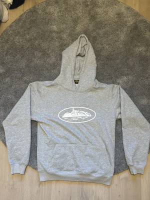 Grå hoodie cortiz - Säljer en grå hoodie från cortiz i storlek medium. Hoodien har ett vitt tryck med motiv på bröstet, stor huva och klassisk magficka. Tillverkad i mjukt bomullsmaterial och har ribbade muddar vid ärmar och nederkant. Perfekt för en chill och avslappnad stil.