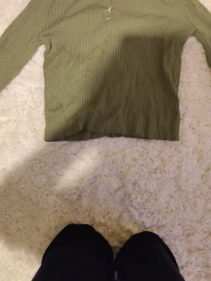 Olivgrön ribbad tröja med dragkedja - Snygg olivgrön ribbad tröja med lång ärm och halv hög krage. Dragkedja framtill med rund metallring som detalj. Perfekt till jeans eller kjol för en trendig look.