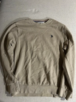 Beige sweatshirt från U.S. Polo Assn. - Stilren beige sweatshirt från U.S. Polo Assn. med rund halsringning och diskret broderad logga på bröstet. Tröjan har långa ärmar och ribbade muddar vid ärmslut och nederkant. Perfekt för en avslappnad och snygg look.