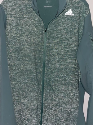 Grön zip-jacka från Adidas - Snygg grön zip-jacka från Adidas med melerad framsida och ryggparti. Jackan har hel dragkedja, hög krage och diskret ficka med dragkedja på ärmen. Adidas-logga i vitt på bröstet. Perfekt för träning eller som sportig vardagsstil.