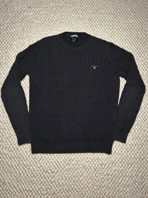 Mörkblå kabelstickad tröja Gant - Snygg mörkblå kabelstickad tröja från Gant med rund halsringning och diskret broderad logga på bröstet. Tröjan har långa ärmar, ribbade muddar och är tillverkad i mjukt material som passar perfekt till vår och höst. Storlek L passar dock M/L.