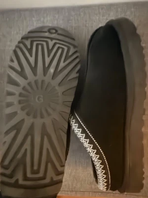 Svarta UGG Slides Tasman - Säljer ett par svarta UGG tofflor i mocka med vit zigzag-mönstrad kant runt öppningen. Tofflorna har en robust gummisula med tydligt mönster och är fodrade för extra komfort. Perfekta för att hålla fötterna varma hemma. Snabb affär-700kr, finns att hämta på Östermalm - 700kr 