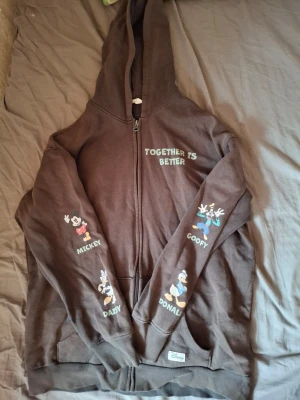 Brun Disney hoodie med tryck - Säljer en brun hoodie med dragkedja från Disney. På framsidan står det 'TOGETHER IS BETTER' och på ärmarna finns tryck av Mickey, Goofy, Daisy och Donald. Baksidan har stort tryck av Piff & Puff och texten 'YOU'RE NUTS!'. Perfekt för dig som älskar klassiska Disney-karaktärer. Har även små färg fläckar på sig för att see kolla dom två sista bilderna 