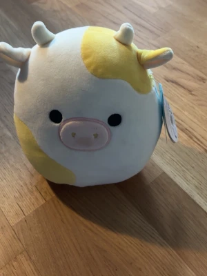 Squishmallows kossa Bobbie gul/vit - Supermjuk och rund gosedjurskossa från Squishmallows, modellen Bobbie. Den har vita och gula fält, små horn och öron samt ett sött broderat ansikte med rosa mule. Perfekt att krama eller ha som dekoration i rummet.