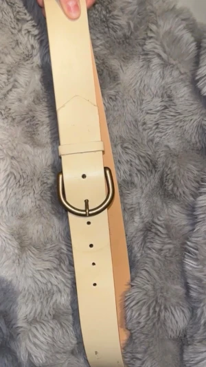 Beige skärp med metallspänne - Stilrent beige bälte med ett robust, rundat metallspänne i guldton. Bältet är brett och tillverkat i slätt skinn, perfekt för att ge en snygg touch till jeans eller klänning. Enkel design utan mönster eller logga.