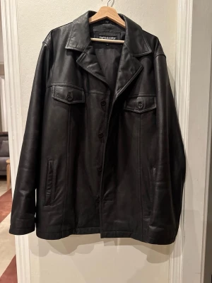 Svart skinnjacka från Steve Madden - Stilren svart skinnjacka från Steve Madden med klassisk krage, knäppning framtill och två bröstfickor med lock och knapp. Jackan har raka ärmar och en tidlös design som passar till många olika looks.