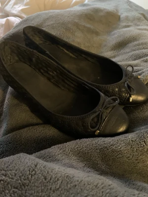  ballerinaskor - Säljer ett par svarta ballerinaskor med quiltat mönster och liten rosett framtill. Skorna har rundad tå och detaljer i guld på rosetten. Materialet ser ut att vara konstläder. Perfekta för dig som gillar klassisk och stilren look.