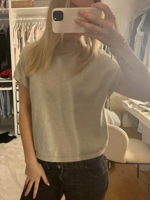 Beige stickad topp med korta ärmar - Säljer en stilren beige stickad topp med korta ärmar och ribbad hög krage. Toppen har ribbade detaljer vid ärmslut och nederkant. Perfekt att matcha med jeans eller kjol för en avslappnad look.