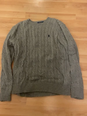 Grå kabelstickad tröja Polo Ralph Lauren - Kabelstickad grå tröja från Polo Ralph Lauren med rund halsringning och ribbade muddar. Liten mörkblå logga broderad på bröstet. Klassisk och tidlös design i mjukt material. 