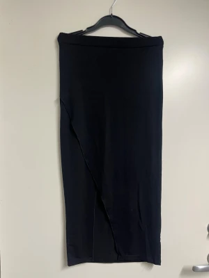 Svart långkjol i enkel design - En stilren svart långkjol med rak passform och minimalistisk look. Den har en elegant slits som överlappar kjolen. Kjolen är gjord i ett mjukt material som faller snyggt och passar perfekt till olika outfits. 